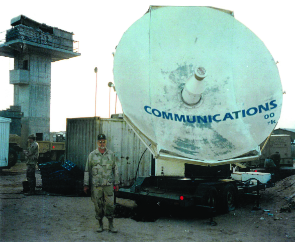 A large, mobile satellite dish with COMMUNICATIONS printed on it, part of a field-deployed communications hub next to a guard tower. A man in desert camouflage stands in front of the dish.